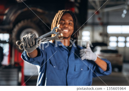 Positive facial expression, holding wrenches. Male mechanic is with car in garage Positive facial expression, holding wrenches. Male mechanic is with car in garage 135605964