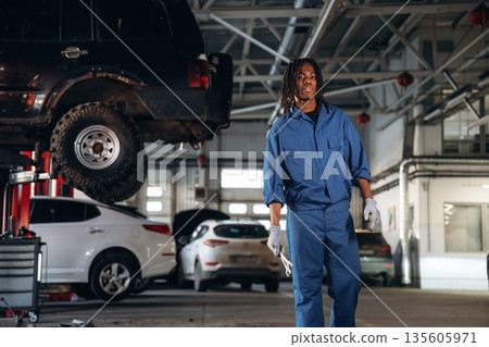 Dreadlocks hairstyle, in blue uniform. Male mechanic is with car in garage Dreadlocks hairstyle, in blue uniform. Male mechanic is with car in garage 135605971
