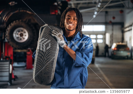 Tire is in hands. Male mechanic is with car in garage Tire is in hands. Male mechanic is with car in garage 135605977