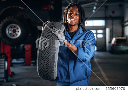 Tire is in hands. Male mechanic is with car in garage Tire is in hands. Male mechanic is with car in garage 135605978