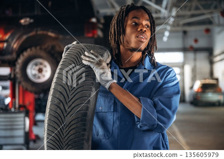 Tire is in hands. Male mechanic is with car in garage Tire is in hands. Male mechanic is with car in garage 135605979