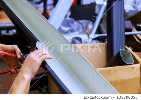 Contractor uses tools to cut metal gutter channels while surrounded by various materials in workspace. 135606055