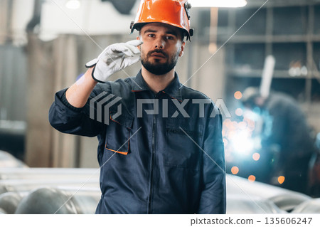 Against blurred background. Welder is standing in protective hat. Man is working in the factory Against blurred background. Welder is standing in protective hat. Man is working in the factory 135606247