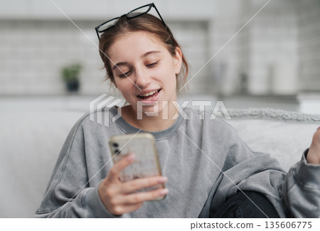 Smiling Young Woman Chatting Via Video Call While Sitting On A Sofa In A Light Room 135606775
