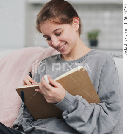 Pretty Girl Sitting On The Sofa At Home Writing Plans And A To-Do List In A Notebook Pretty Girl Sitting On The Sofa At Home Writing Plans And A To-Do List In A Notebook 135606776
