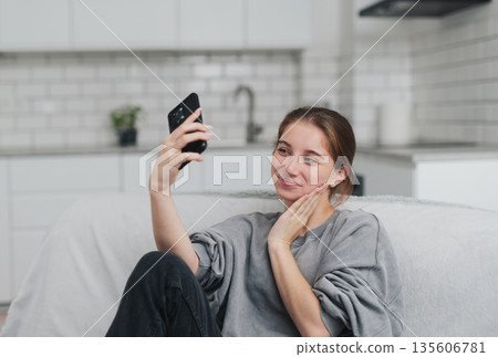 Happy, Pretty Young Girl Sitting On The Sofa At Home, Taking A Selfie On Smartphone Happy, Pretty Young Girl Sitting On The Sofa At Home, Taking A Selfie On Smartphone 135606781
