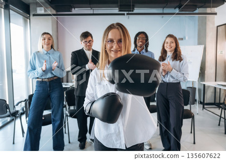 Woman is in boxing gloves. Group of young business people are in conference room Woman is in boxing gloves. Group of young business people are in conference room 135607622