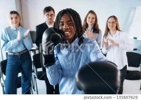 Black man is with boxing gloves. Group of young business people are in conference room 135607629