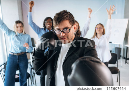Punching, in boxing gloves. Group of young business people are in conference room Punching, in boxing gloves. Group of young business people are in conference room 135607634