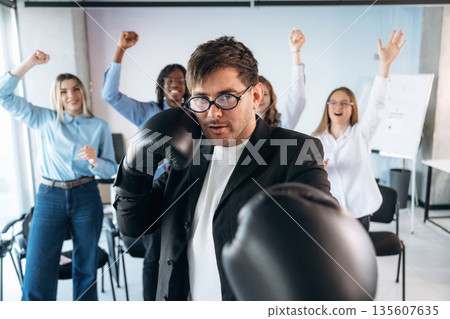 Focused guy in glasses is with boxing gloves. Group of young business people are in conference room Focused guy in glasses is with boxing gloves. Group of young business people are in conference room 135607635