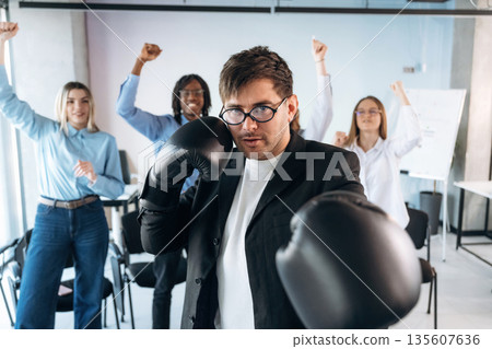 Focused guy in glasses is with boxing gloves. Group of young business people are in conference room Focused guy in glasses is with boxing gloves. Group of young business people are in conference room 135607636