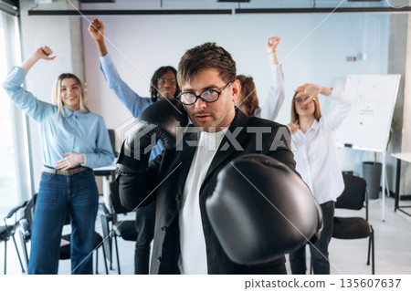 Focused guy in glasses is with boxing gloves. Group of young business people are in conference room 135607637
