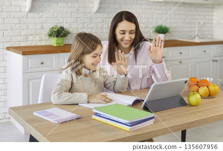 Mother and daughter with tablet happily waving during video call at kitchen table 135607956