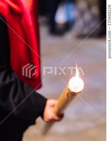 Close up of a person in a red hood holding a lit wax candle at night. 135608226