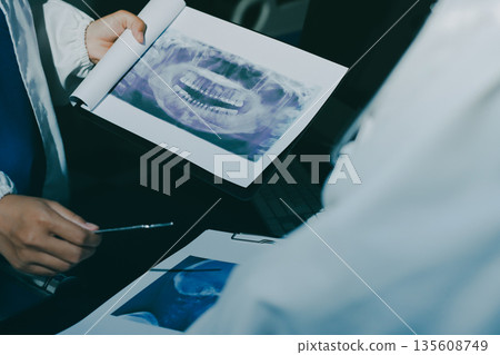 Two doctors and a female nurse meet at a table in the hospital, collaborating on medical tasks using laptops and computers Two doctors and a female nurse meet at a table in the hospital, collaborating on medical tasks using laptops and computers 135608749
