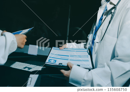 Two doctors and a female nurse meet at a table in the hospital, collaborating on medical tasks using laptops and computers Two doctors and a female nurse meet at a table in the hospital, collaborating on medical tasks using laptops and computers 135608750