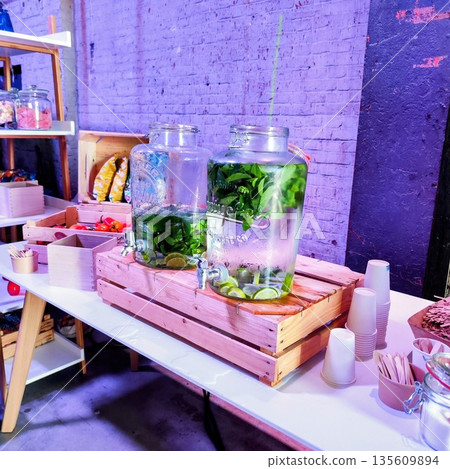 Two large glass jars filled with refreshing beverages are displayed on a table. Each jar contains water infused with lime slices and fresh mint leaves, resting on wooden stands with spigots 135609894