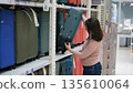 Young woman examining a travel suitcase in a luggage aisle, checking wheels and handle for comfort and durability, travel preparation and vacation planning 135610064