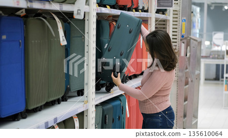 Young woman examining a travel suitcase in a luggage aisle, checking wheels and handle for comfort and durability, travel preparation and vacation planning Young woman examining a travel suitcase in a luggage aisle, checking wheels and handle for comfort and durability, travel preparation and vacation planning 135610064