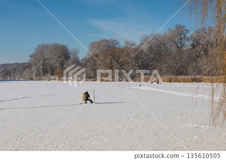 A fisherman sits on a snow-covered frozen river on a clear day A fisherman sits on a snow-covered frozen river on a clear day 135610505