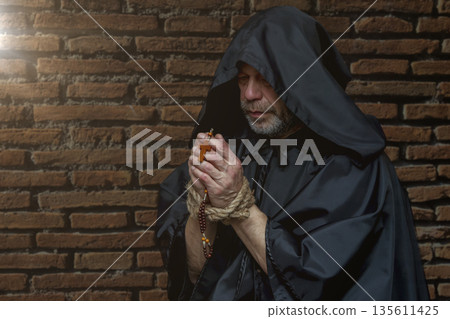 Monk in dark clothing holds rosary with bound hands against old brick wall. 135611425