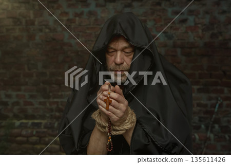 Monk in hood looks straight at camera holding rosary with bound hands. 135611426