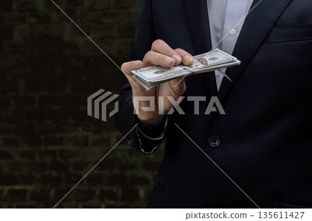 A businessman in a formal suit holding a stack of US dollars against a brick wall 135611427