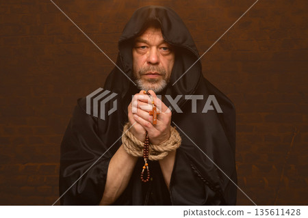 Monk in dark hood holds rosary and prays with bound hands near wall. 135611428