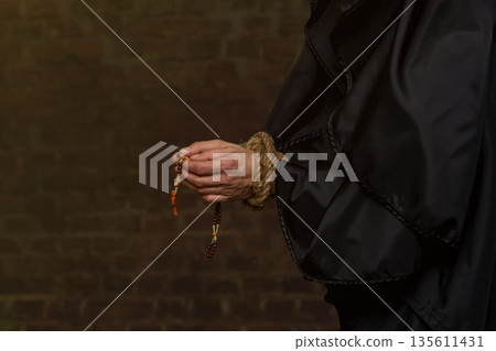 Close-up of a monk's hands tied with rope, holding rosary beads against a blurred dark background. 135611431