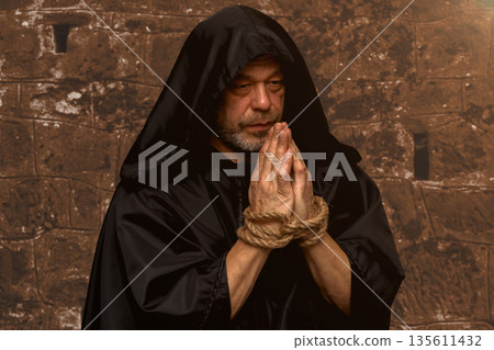 An elderly monk in a black hood stands with his hands tied and prays with a rosary against a brick wall. 135611432