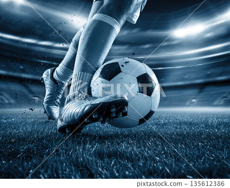Soccer player kicks ball during match at night in stadium with bright lights shining Soccer player kicks ball during match at night in stadium with bright lights shining 135612386