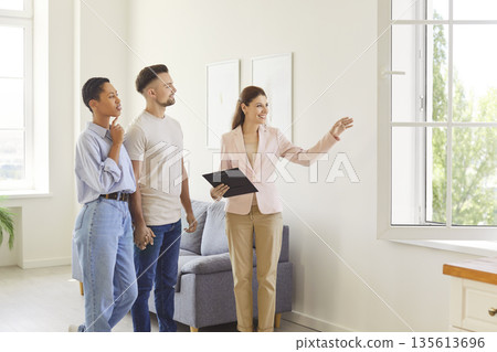 Female realtor showing house to a young happy couple looking at the view from the window. 135613696