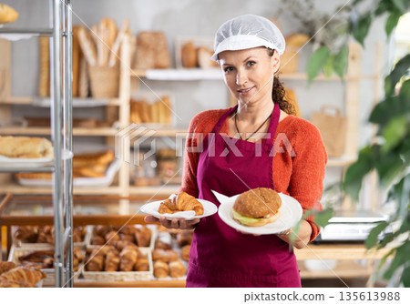 Woman entrepreneur, owner of bakery, holds out sandwich in disposable dish plate in hand Woman entrepreneur, owner of bakery, holds out sandwich in disposable dish plate in hand 135613988