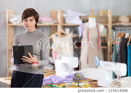 Female tailor draws sketch of future clothing on an electronic tablet. Working with modern gadgets in sewing workshop 135614066