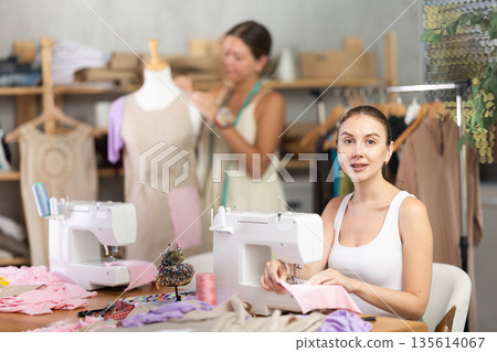 Woman seamstress operating by sewing machine. Assistant work with mannequin in background Woman seamstress operating by sewing machine. Assistant work with mannequin in background 135614067