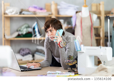 Woman tailor worker talks to customer on phone, laptop on table Woman tailor worker talks to customer on phone, laptop on table 135614100