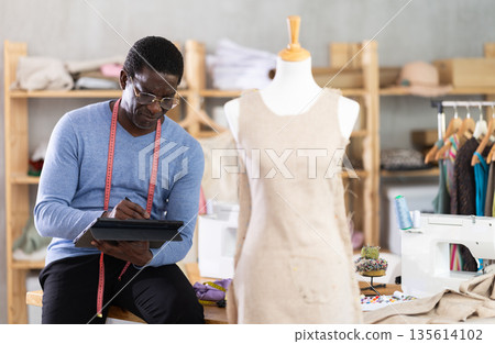 Male tailor draws sketch of future clothing on an electronic tablet. Working with modern gadgets in sewing workshop 135614102