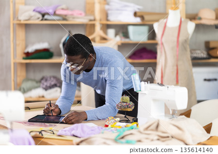 African American man tailor creates sketch of dress on tablet African American man tailor creates sketch of dress on tablet 135614140