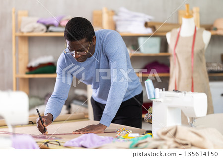 Male tailor draws sketch of future clothing on paper using patterns in sewing workshop 135614150