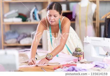 Attentive young female dressmaker drawing on textile in sewing workshop 135614159