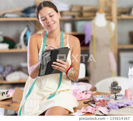 Young female dressmaker drawing on tablet in sewing workshop 135614167