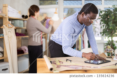 Male tailor draws sketch of future clothing on an electronic tablet. Working with modern gadgets in sewing workshop 135614236