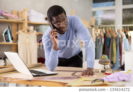 African man tailor worker talks to customer on phone, laptop on table 135614272