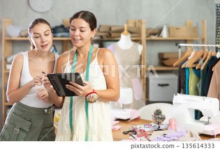Two female dressmakers drawing a model of clothes on a tablet 135614333