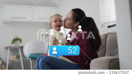 Mother holding baby, leaning in kissing her cheek in living room, with blue notification showing 52 135617307