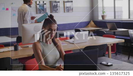Typing teenage girl working at laptop in modern shared office, with co-workers reviewing documents 135617325