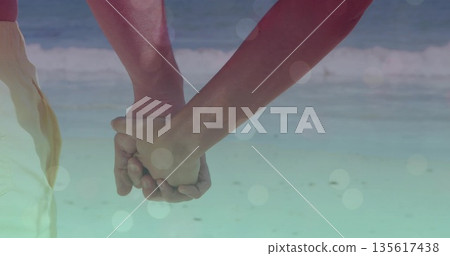 Holding hands of couple wearing light beach shorts, pink sleeveless top in shallow surf, soft flare 135617438