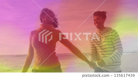 Walking couple wearing mustard cable-knit turtleneck and striped hoodie on sandy shoreline, smiling 135617439