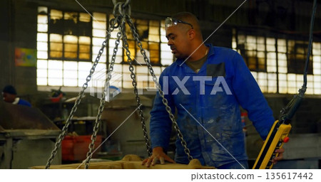 Operating chain hoist lifting casting at plant, worker in coverall using yellow remote, copy space 135617442