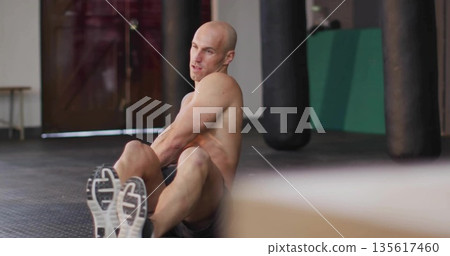 Twisting shirtless athlete performing core exercise on gym floor, with punching bags, glass doors 135617460